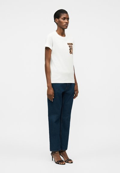 Γυναίκα που φοράει λευκό μπλουζάκι με λογότυπο αρκουδάκι, σκουρόχρωμο μπλε τζιν και μαύρα σανδάλια με ψηλό τακούνι, στέκεται μπροστά σε ένα απλό λευκό φόντο.