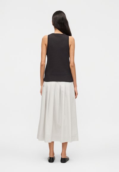 Γυναίκα με μακριά σκούρα μαλλιά που φοράει αμάνικο μαύρο τοπ, ανοιχτό γκρι πλισέ midi φούστα και μαύρα παπούτσια μπαλέτου, στέκεται με την πλάτη προς τα πίσω.