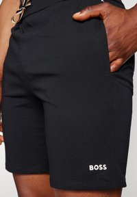 Mustat shortsit, jotka on valmistettu venyvästä kankaasta, sisältävät sivutaskun ja valkoisen "BOSS"-logon alahaarassa. Säädettävä vyötärö näkyy yksityiskohtana.