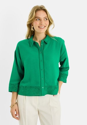 Mujer sonriente, vestida con una blusa abotonada de color verde brillante con detalles de encaje en el cuello, puños y dobladillo, combinada con pantalones beige claro.