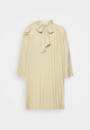 Robe plissée couleur crème avec manches longues et un grand nœud noué au niveau de l'encolure sur un fond uni clair.