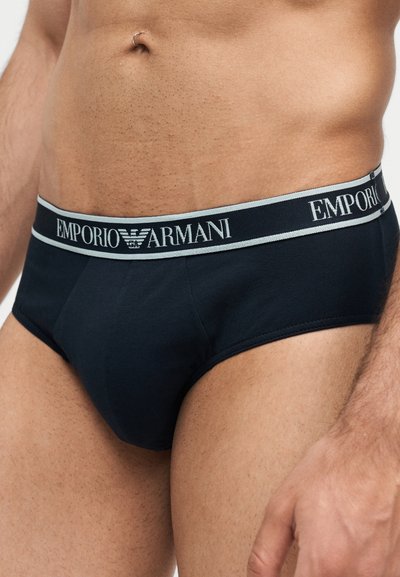 Αρσενικός κορμός με μαύρα εσώρουχα Emporio Armani με λογότυπο στη μέση, που δείχνει τρυπημένο ομφαλό και μέρος του δεξιού χεριού στο πλάι.