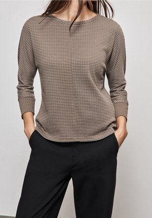 Haut texturé marron avec un motif à carreaux, col rond, manches trois-quarts et ourlet légèrement courbé. Associé à un pantalon noir.