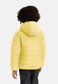 Chaqueta acolchada amarilla con capucha, diseño acolchado y textura acanalada. Presenta un acento reflectante cerca del dobladillo. Llevada sobre pantalones negros.