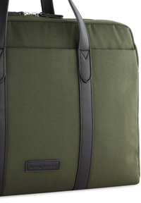 Oliven grøn stoftaske med sorte læderdetaljer, med et robust design, blød tekstur og et logo-patch foran.
