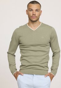 Pull à col en V vert olive en tissu doux, avec un col blanc contrastant et des poignets et un ourlet côtelés. Design ajusté.