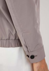 Giacca bomber grigia chiara con una texture liscia e lucida, vita elastica e polsini con bottoni a pressione e un dettaglio con un unico bottone nero.