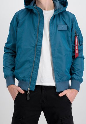 Personne portant une veste à capuche turquoise avec une étiquette rouge "Retirer avant le vol" sur la manche, un t-shirt blanc et un pantalon noir, les mains dans les poches.