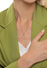 Rosenrødt halskæde med en cirkulær vedhæng på en fin kæde, båret over en hvid top mod en tekstureret grøn blazer.