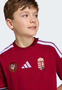 Červený dres s krátkym rukávom a bielymi pruhmi, s vyšívaným erbom Maďarska a logom. Vyrobený z látky s hladkou textúrou.