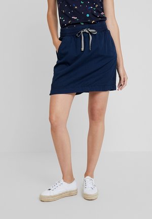 Woman wearing a navy blue drawstring skirt, patterned top, and white platform sneakers standing with one hand in pocket.