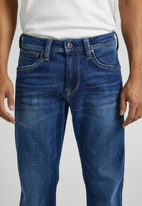 Vaqueros de denim azul con corte recto, que presentan un clásico diseño de cinco bolsillos, desvanecimiento ligero, costuras amarillas y cierre de botón de latón.