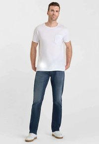 T-shirt blanc à manches courtes avec une poche poitrine, associé à un jean bleu et des baskets blanches. Le jean présente une coupe classique et un design délavé.