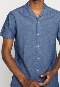 Camicia a maniche corte con bottoni in misto lino blu, presenta un colletto da campeggio, chiusura con bottoni e un tessuto testurizzato con leggere sfumature.