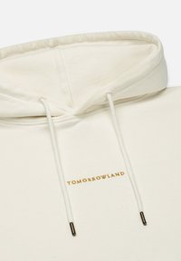 Cremfarbener Kapuzenpullover mit Kordelzug, ausgestattet mit einem goldenen "TOMORROWLAND"-Logo. Weicher Stoff und minimalistisches Design, das eine einfache Konstruktion zeigt.