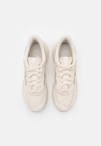 Beige sportskor med texturerat tyg, snörning och en vadderad sula. Sidan har en diskret reflekterande rand. Platta snören.