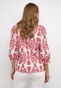 Rosa Bluse mit roten gestickten Blumen- und geometrischen Mustern. Hat Dreiviertelärmel und geraffte Akzente an den Manschetten.
