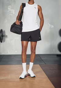 Vit ärmlös topp, svarta shorts med logotyp, vita strumpor och vita sportskor med svarta detaljer. Håller en svart gymväska.