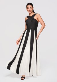Femme portant une robe maxi plissée noire et blanche sans manches avec un décolleté croisé, souriante et posant sur un fond uni.