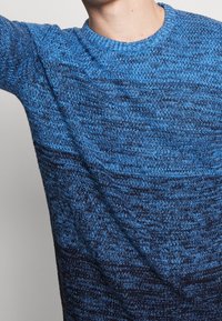 Maglione lavorato a maglia blu con un motivo testurizzato, scollo rotondo e maniche lunghe, che passa da tonalità di blu chiaro a blu scuro.