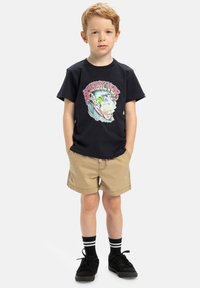 Svart bomullströja med färggrafik av vågor, tillsammans med bruna shorts. Barnet har på sig svarta skor och randiga svartvita strumpor.