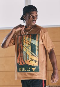 Smeđa pamučna majica s grafičkim dizajnom drveća i tekstom "Chicago Bulls" u podebljanim, kontrastnim bojama, upotpunjena zlatnim lančićem.