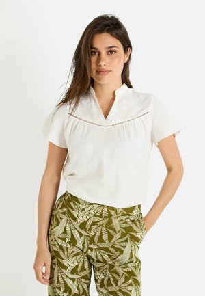 Blouse blanche à manches courtes avec un col festonné, ornée de broderies florales et de découpes au niveau de la poitrine, associée à un pantalon vert à motifs.