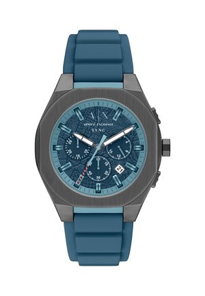 Hodinky s modrým silikonovým řemínkem, texturovaným modrým ciferníkem, stříbrnými detaily, chronografovými subciferníky a zobrazením data na pozici 6 hodin. Osmihranné pouzdro.