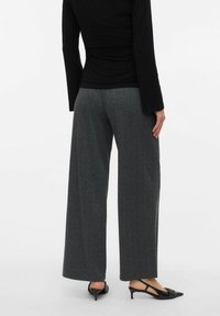 Personne portant un pantalon large gris foncé, un haut noir à manches longues et des escarpins à talons kitten noirs, se tenant devant un fond uni.