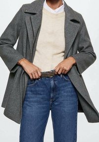 Persona que lleva un abrigo de lana gris sobre un suéter beige y una camisa blanca, abrochándose un cinturón marrón en unos vaqueros azules.