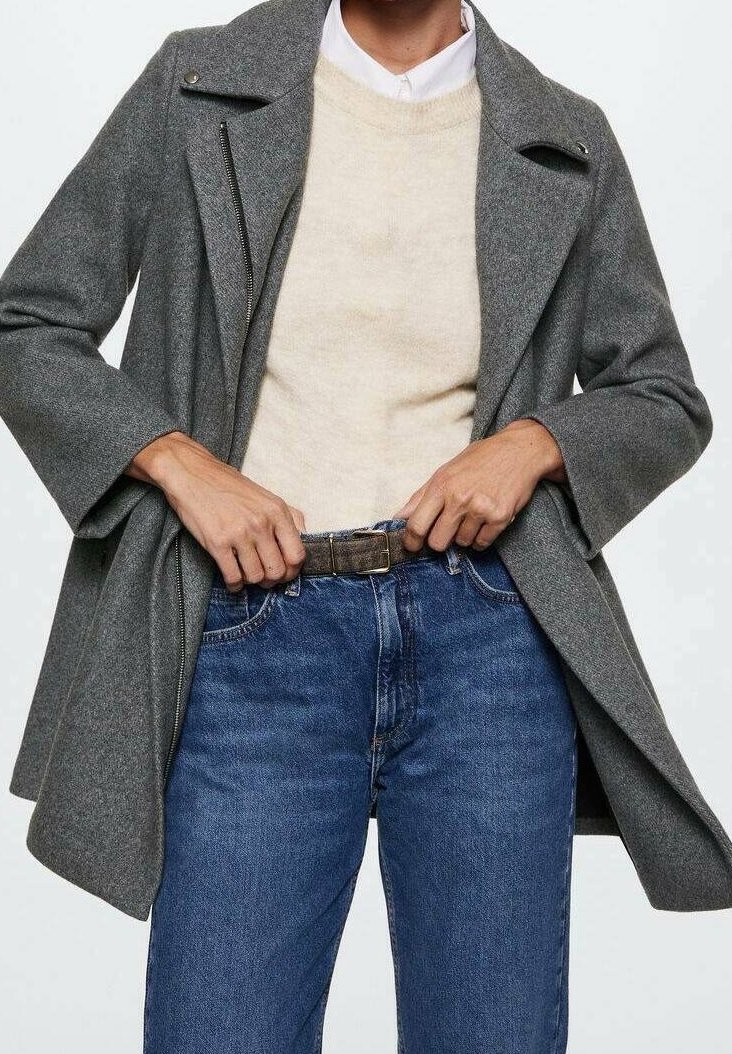 Persona que lleva un abrigo de lana gris sobre un suéter beige y una camisa blanca, abrochándose un cinturón marrón en unos vaqueros azules.