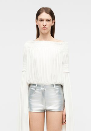 Γυναίκα με ίσια καστανά μαλλιά που φοράει λευκή μπλούζα χωρίς ώμους με μακριά μανίκια και μεταλλικά ασημί σορτς με ψηλή μέση.