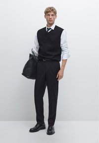 Man in black sleeveless sweater vest, white shirt with rolled sleeves, black tie, black trousers, and black shoes holding black leather bag.
