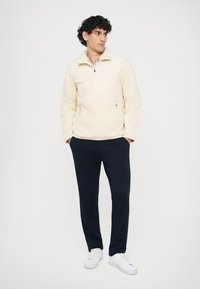 Pullover di pile crema con colletto alto, zip a metà e tasca frontale; abbinato a pantaloni della tuta blu scuro e scarpe da ginnastica bianche.