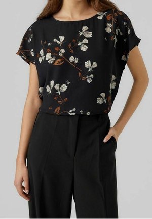 Kobieta ubrana w czarną bluzkę z krótkim rękawem z białym i brązowym kwiatowym wzorem, połączoną z czarnymi spodniami z wysokim stanem, ręka w kieszeni.