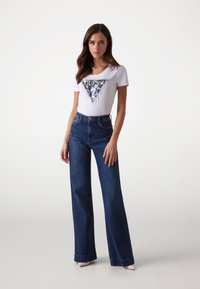 Hvid t-shirt med et blåt trekantsgrafiklogo, parret med højtaljede mørkeblå brede jeans. Glat stof, afslappet design.