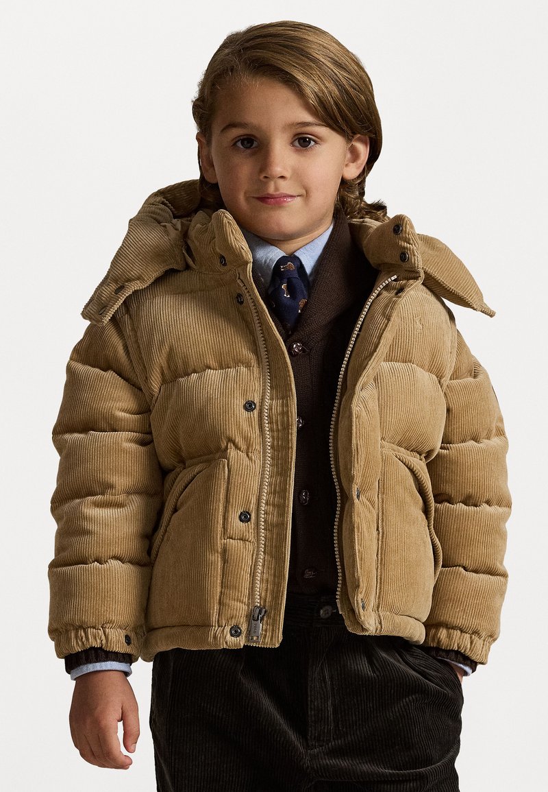 Παιδικό μπουφάν με καλυμμένο χουτιά σε μπεζ χρώμα, με αποσπώμενη κουκούλα, μπροστινές τσέπες και μαύρους κουμπάκους. Συνδυασμένο με ένα καφέ γιλέκο και μπλε πουκάμισο.