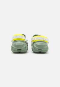 Green slip-on shoes with textured rubber soles, featuring adjustable yellow straps and white hardware. Rear view showcases streamlined design.