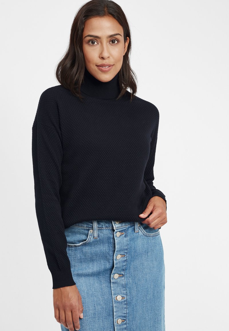 Dunkelblauer Rollkragenpullover mit strukturiertem Strickdesign, kombiniert mit einem hellblauen Denim-Rock mit Knopfleiste. Klare, definierte Formen und Farben.