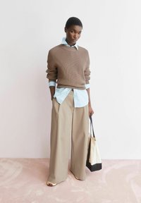 Persoon staat en draagt een bruine gebreide trui over een blauw overhemd, beige wijde broekspijpen, sneakers, en houdt een zwart-witte draagtas vast.