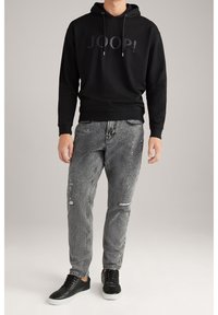 Sudadera negra con capucha y logo, jeans grises desgastados con salpicaduras de pintura y rasgaduras, y zapatillas negras con suelas blancas.