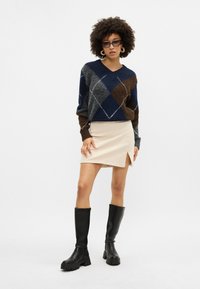 Sweter w miękkim dzianinie w wzór argyle w odcieniach granatu, brązu i szarości; beżowa mini spódnica z bocznym rozcięciem; czarne kozaki do kolan na masywnych podeszwach.