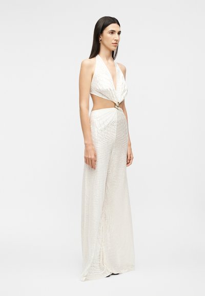 Γυναίκα όρθια, φορώντας λευκό φόρεμα με λαιμόκοψη halter, πλαϊνά ανοίγματα και υφασματικό, λαμπερό σχέδιο, με το προφίλ της προς τα πλάγια πάνω σε απλό φόντο.