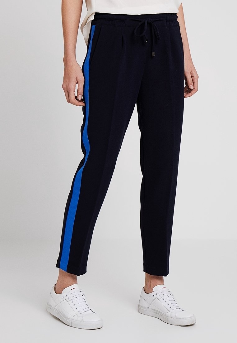 Personne portant un pantalon de jogging noir avec des bandes latérales bleues et des baskets blanches, debout devant un fond gris clair.