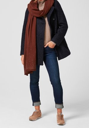 Manteau texturé bleu marine, écharpe marron avec accents métalliques, haut beige clair, jean denim foncé et baskets beige clair avec lacets et logo.