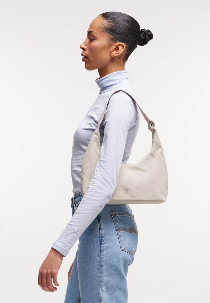 Woman with hair in bun wearing a light turtleneck and jeans carrying a beige shoulder bag against a plain white background.