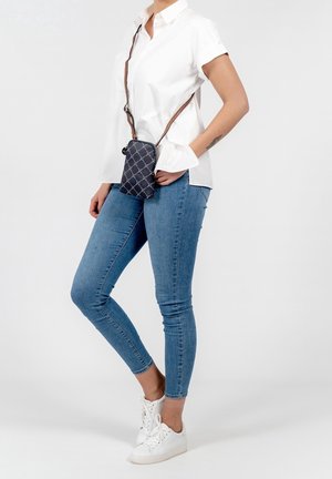 Borsa a tracolla blu con un motivo geometrico, tracolla marrone, forma rettangolare compatta, chiusura con zip, indossata sopra una camicia bianca e jeans blu.
