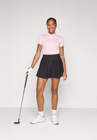 Mosolygó nő rózsaszín pólót és fekete, pliszírozott golf szoknyát visel, kezében golfütő, fehér tornacipő és kesztyű van rajta. Egyszerű szürke háttér.
