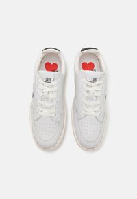 White leather sneakers with a rounded toe, perforated detailing, flat white laces, and a solid black heel accent; red heart design on insoles.