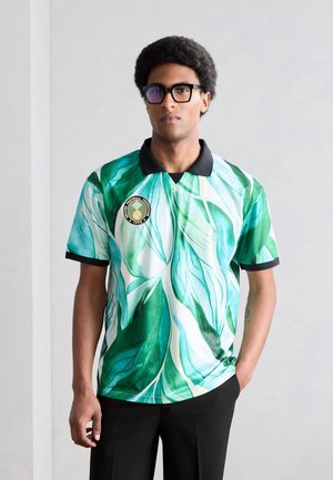 Férfi fekete szemüvegben és fekete nadrágban, zöld-fehér levélmintás fekete galléros pólóingben, áll egy kézzel a zsebében.